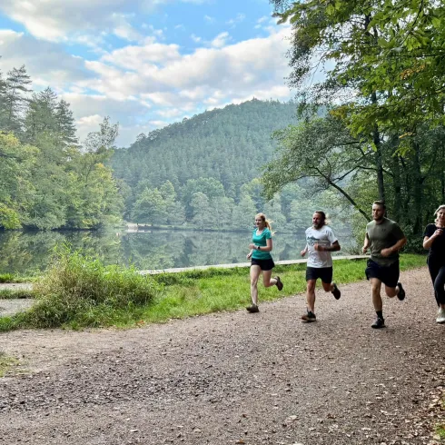 Eine Gruppe von vier Personen joggt entlang eines malerischen Weges am Wasser. Im Hintergrund sind Bäume und eine hügelige Landschaft zu sehen.