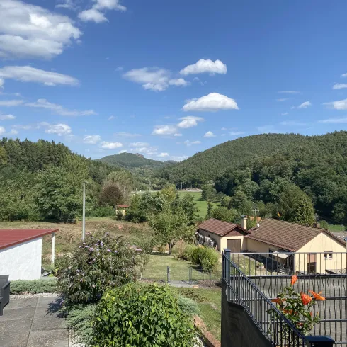 Eine malerische Landschaft mit sanften Hügeln und grünem Wald. Im Vordergrund sind einige Häuser und eine Terrasse zu sehen.