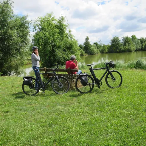 Zwei Personen genießen die Natur an einem ruhigen See. Neben ihnen stehen Fahrräder auf dem grünen Gras.
