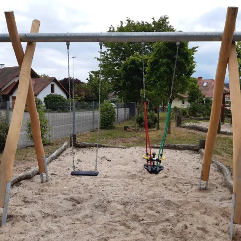 Ein Spielplatz mit einer Schaukel in einem Sandbereich. Im Hintergrund sind Bäume und einige Häuser sichtbar.