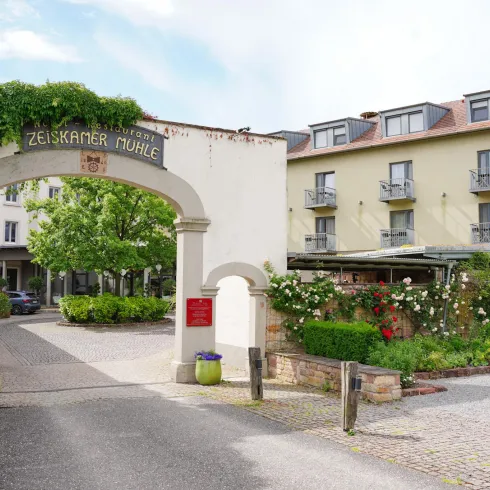 Ein einladender Eingang zu einem Hotel mit einem blühenden Garten. Das Gebäude ist umgeben von Bäumen und bietet eine entspannte Atmosphäre.