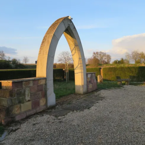 Ein eleganter Bogen aus Stein steht auf einem Kiesweg. Im Hintergrund sind sanfte Hügel und Bäume zu sehen.