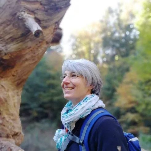 Eine Frau mit grauen Haaren steht in einem Wald und lächelt. Im Hintergrund sind Bäume mit herbstlichen Farben zu sehen.
