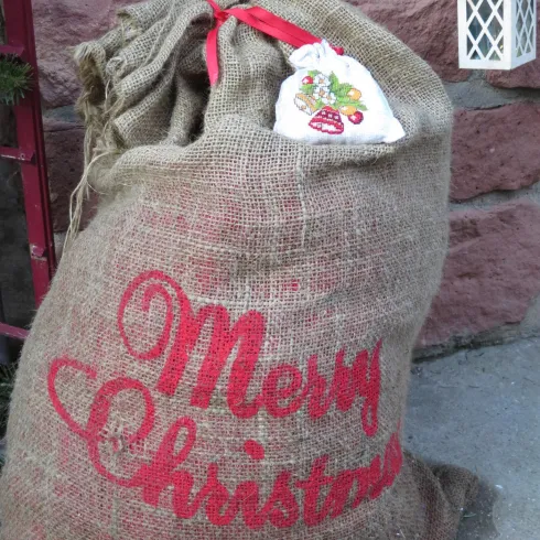 Ein großer Jutesack mit der Aufschrift "Merry Christmas". Der Sack ist festlich dekoriert und hat eine rote Schleife.
