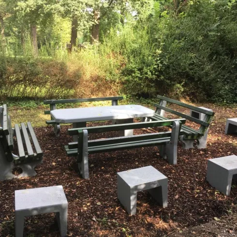 Ein rustikaler Tisch mit Bänken und Hockern in einem ruhigen Garten. Umgeben von Bäumen und Sträuchern.