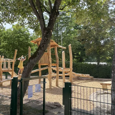 Ein Spielplatz aus Holz mit verschiedenen Kletterelementen und Rutsche. Die Umgebung ist grün und von Bäumen umgeben.