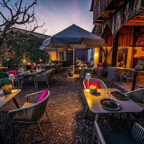 Ein gemütlicher Außenbereich eines Restaurants mit modernen Tischen und Stühlen. Die Atmosphäre ist entspannt, umgeben von Pflanzen und einer sanften Beleuchtung.