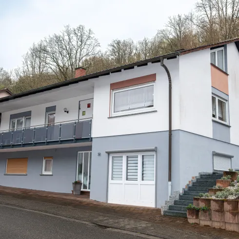 Ein modernes Haus mit einem Balkon und einer hellen Fassade. Im Hintergrund sind Bäume sichtbar, die eine ruhige Umgebung schaffen.