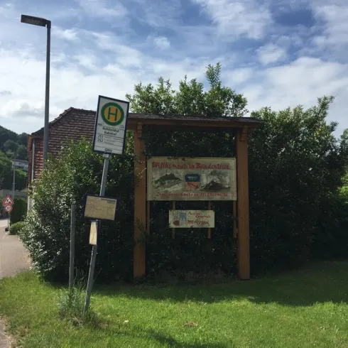 Ein Schild, das ein Restaurant oder eine Gaststätte anzeigt, umgeben von Bäumen und Sträuchern. Im Hintergrund sind leicht hügelige Landschaften und ein klarer Himmel zu sehen.
