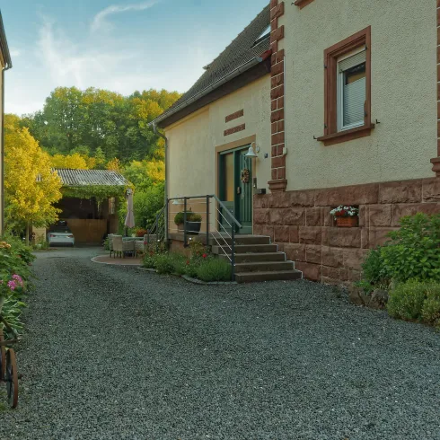 Eine ruhige Einfahrt mit einer gepflasterten Oberfläche und begrünten Bereichen. Auf der rechten Seite steht ein charmantes Gebäude mit einer Treppe und blühenden Pflanzen.
