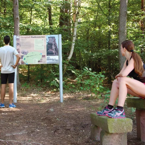 Eine Frau sitzt auf einer Bank im Wald und beobachtet einen Mann, der ein Informationsschild liest. Um sie herum sind viele Bäume und grünes Pflanzenmaterial.