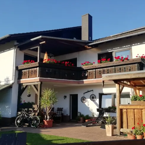 Ein schönes Haus mit einem Balkon, der mit roten Blumen geschmückt ist. Im Vordergrund stehen Pflanzen und Fahrräder.