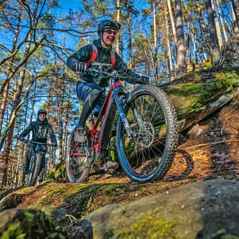 Zwei Mountainbiker fahren durch einen Waldweg. Der vordere Fahrer überwindet steinige Stellen mit viel Spaß.