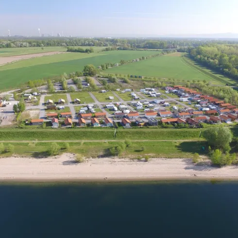 Ein schöner Campingplatz am Wasser, umgeben von grünen Wiesen und Bäumen. Im Vordergrund ist ein sandiger Strand und dahinter mehrere Stellplätze mit Wohnmobilen und Ferienhäusern zu sehen.