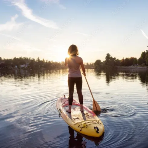 Eine Person steht auf einem Paddleboard auf ruhigem Wasser und paddelt. Der Sonnenuntergang spiegelt sich im Wasser und sorgt für eine entspannte Stimmung.