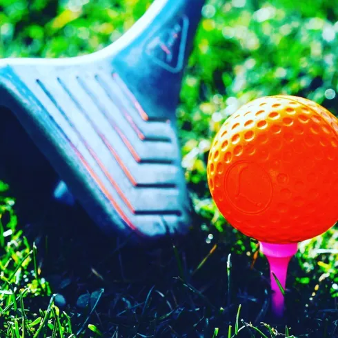Ein Golfschläger liegt im Gras neben einem orangefarbenen Golfball auf einem Tee. Die Szene ist hell und einladend.