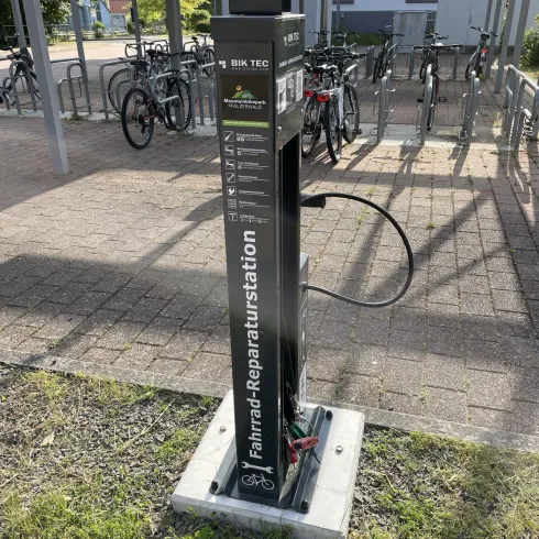 Eine Fahrrad-Reparaturstation mit verschiedenen Werkzeugen steht an einem Fahrradparkplatz. Im Hintergrund sind mehrere Fahrräder abgestellt.