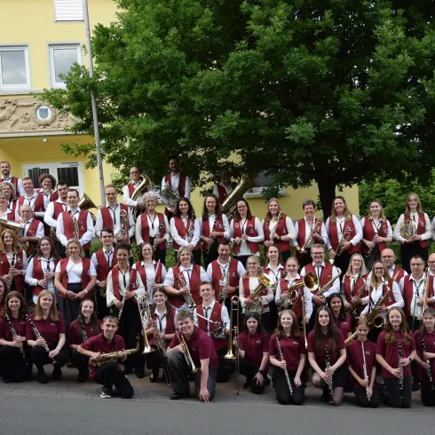 Eine große Musikgruppe steht vor einem gelben Gebäude. Die Mitglieder tragen einheitliche Kleidung in Weinrot und Weiß und halten verschiedene Musikinstrumente.