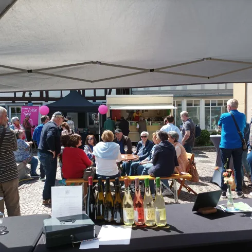 Ein lebhaftes Fest mit vielen Menschen in einer Marktsituation. Stände und Tische sind sichtbar, und die Atmosphäre ist fröhlich und gesellig.