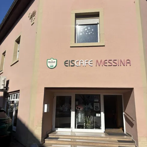Ein Eiscafé namens Messina mit einer einladenden Eingangsfront. Das Gebäude ist in einem hellen Farbton gestrichen und hat große Fenster.