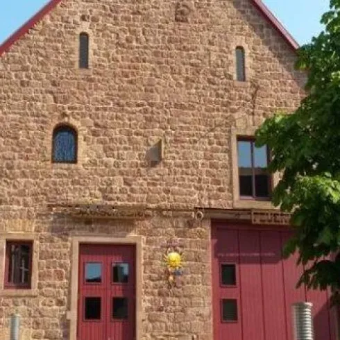Ein altes, steinernes Gebäude mit roten Türen und einem modernen Dach. Vor dem Gebäude steht ein kleiner Baum.