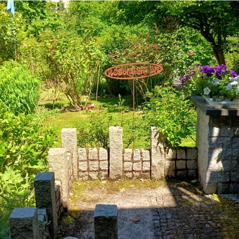 Ein schöner Garten mit üppigem Grün und bunten Blumen. Im Hintergrund steht ein dekoratives Schild und eine kleine Steintreppe führt hinunter.