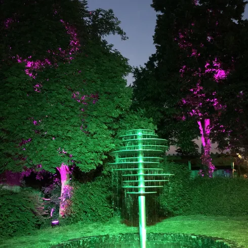 Ein schöner Wasserbrunnen in einem parkähnlichen Garten. Die Bäume sind mit buntem Licht beleuchtet, das eine gemütliche Atmosphäre schafft.