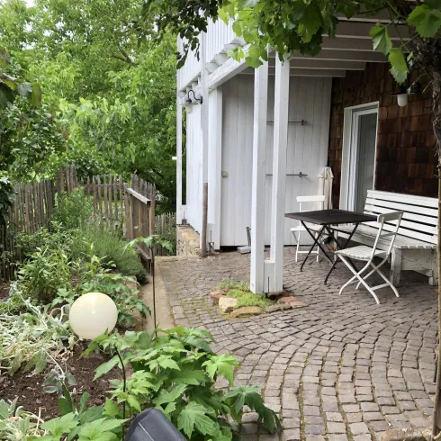 Ein gemütlicher Außenbereich mit einer Terrasse und einem kleinen Tisch. Umgeben von üppigen Pflanzen und einem gepflasterten Weg.