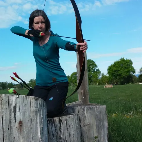 Eine Person schießt mit einem Bogen auf ein Ziel. Im Hintergrund sieht man eine grüne Wiese und einen blauen Himmel.