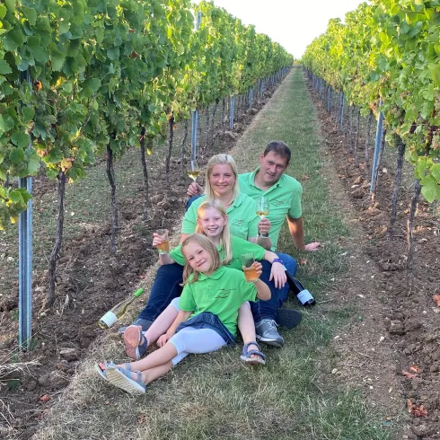 Eine Familie sitzt im Weinberg zwischen den Reben. Alle tragen grüne T-Shirts und lächeln in die Kamera.