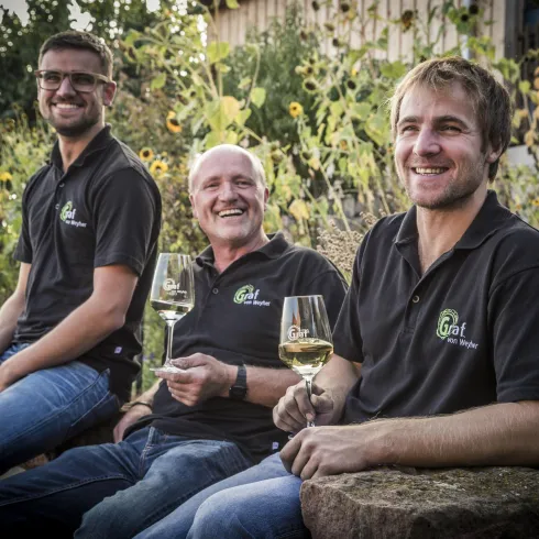 Drei Männer sitzen entspannt im Freien und halten Gläser mit Wein. Im Hintergrund sind Weinreben zu sehen.