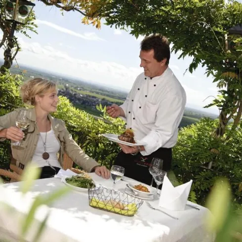 Eine elegante Restaurantatmosphäre im Freien mit einer Person, die ein Glas Wein hält. Ein Kellner serviert ein Gericht in einer malerischen Umgebung.
