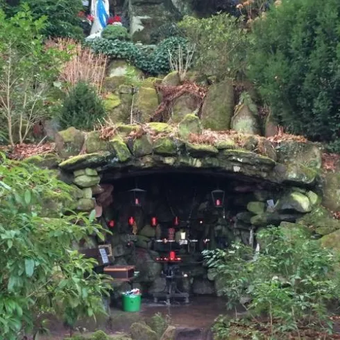 Eine kleine Höhle aus Stein mit Kerzen und religiösen Gegenständen. Darüber steht eine Statue der Jungfrau Maria, umgeben von grüner Vegetation.