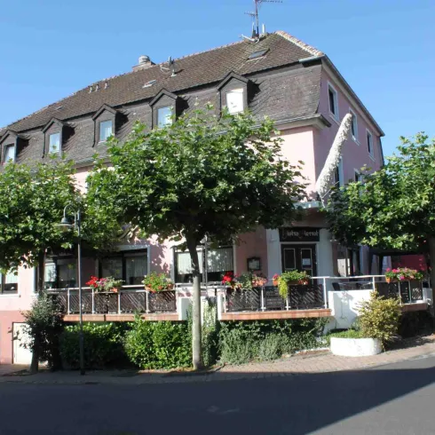 Ein charmantes Gebäude mit einem Balkon, umgeben von Bäumen und Blumen. Die Straße führt sanft nach oben und ist sonnig.