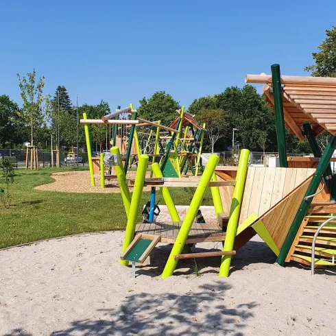 Ein Spielplatz mit bunten Spielgeräten und einer Rutsche steht in einem grünen Park. Der Himmel ist klar blau und es gibt viele Bäume in der Umgebung.