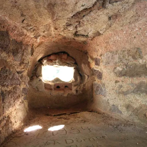 Ein kleiner, dunkler Raum mit Steinmauern und einem Fenster, das Licht hineinlässt. Der Boden ist rau und zeigt einige eingravierte Zeichen.