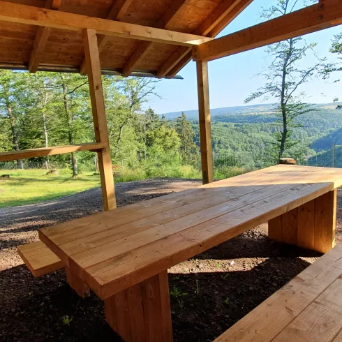 Ein gemütlicher Platz unter einem Holzdach mit einem langen Tisch und Bänken. Im Hintergrund ist eine schöne Aussicht auf die hügelige Landschaft und Bäume.
