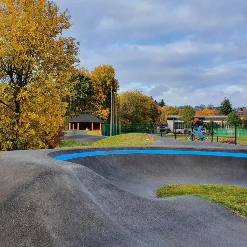 Ein Skatepark mit einer gepflasterten Einrichtung und einer blauen Linie. Im Hintergrund sind Bäume mit Herbstlaub und ein sportlicher Bereich zu sehen.
