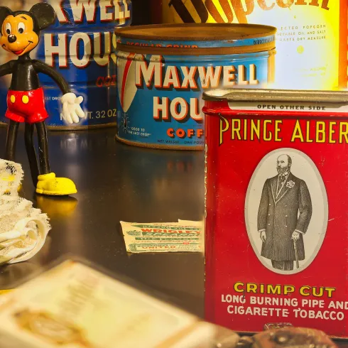 Eine Sammlung von Vintage-Dosen und einem Spielzeug in Form eines Mickey-Mouse-Hundes. Im Vordergrund befindet sich eine rote Dose mit dem Aufdruck "PRINCE ALBERT" für Tabak.