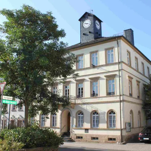 Ein historisches Gebäude mit einer Uhr am Dach. Es befindet sich in einer ruhigen Straßenumgebung mit Bäumen.