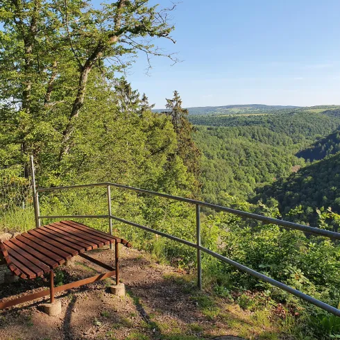 Ein Aussichtspunkt mit einer Holzliege und Blick auf die bewaldete Landschaft. Die Sonne scheint auf einen klaren Himmel, was eine ruhige Atmosphäre schafft.