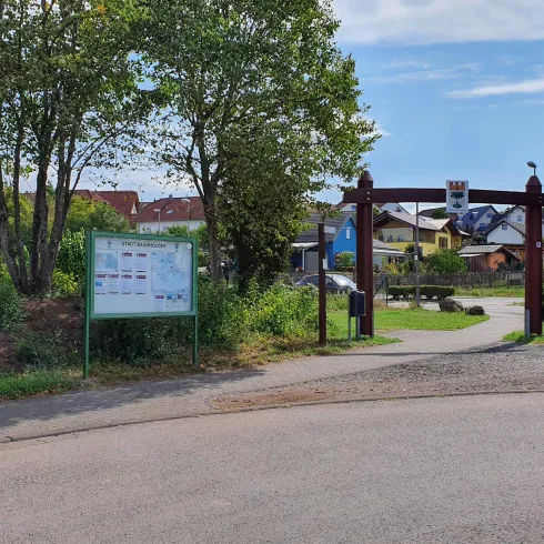 Ein Eingang zu einem Freizeitbereich mit einer hölzernen Pforte und grünen Bäumen. Der Himmel ist klar und die Umgebung wirkt einladend.