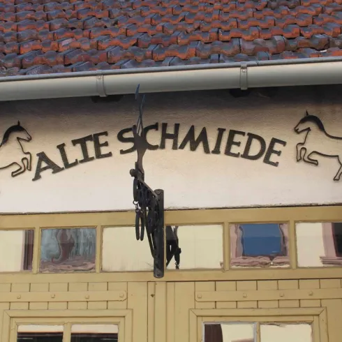 Ein altes Schmiedegebäude mit dem Schriftzug "ALTE SCHMIEDE" über dem Eingang. Um den Schriftzug sind stilisierte Pferde dargestellt.
