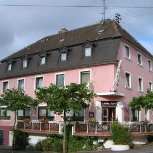 Ein pinkes Hotelgebäude mit mehreren Fenstern und einem Eingang. Umgeben von Bäumen und einer ruhigen Atmosphäre.