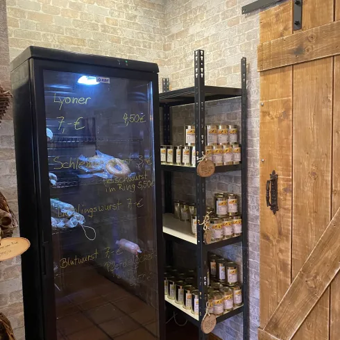 Ein Kühlregal mit verschiedenen Fleischwaren und Konserven steht in einem gemütlichen Laden. Die Wände sind mit Ziegelstein gestaltet und eine Holzschiebetür ist sichtbar.