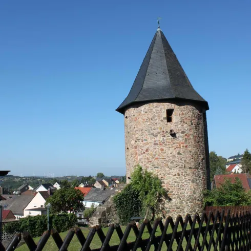 Ein alter runder Turm mit einem spitzen Dach steht in einer malerischen Landschaft. Im Hintergrund sind Häuser und Bäume sichtbar.