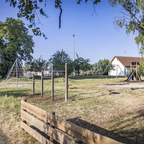 Ein Spielplatz mit Schaukeln und Klettergerüst in einem offenen, grünen Bereich. Im Hintergrund ist ein kleines Gebäude zu sehen, umgeben von Bäumen.