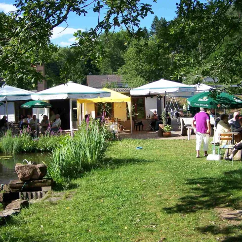 Ein gemütliches Café im Freien mit mehreren Schirmen und Tischen. Umgeben von grünem Gras und Bäumen, genießen die Gäste die sonnige Atmosphäre.