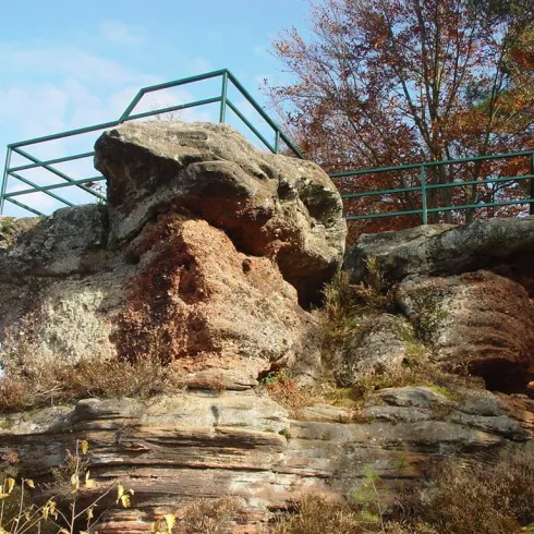 Ein großer, rauer Felsen mit einer grünen Aussichtsplattform. Umgeben von buntem Laub und einem klaren Himmel.