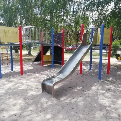 Ein Spielplatz mit einer großen Rutsche und bunten Spielgeräten. Die Umgebung ist von Bäumen umgeben und der Boden ist mit Sand bedeckt.